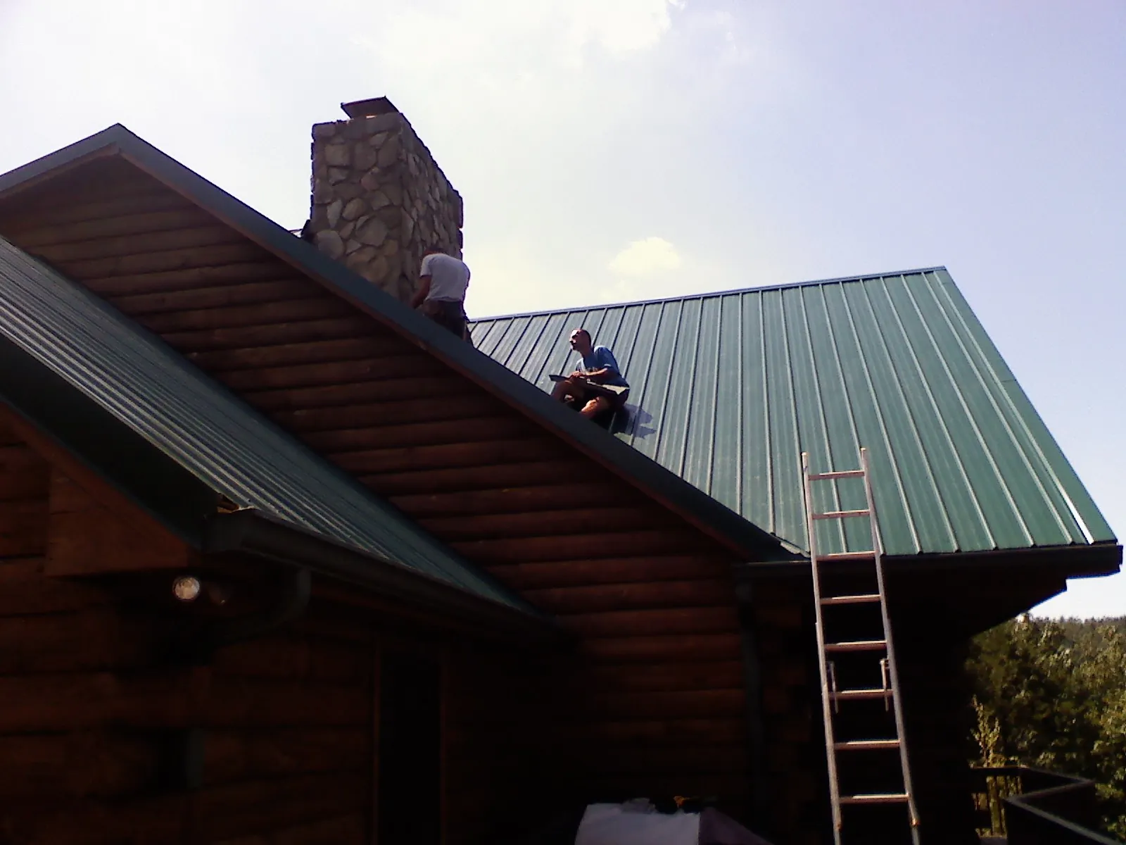 Roofers installing green metal panels beside a stone chimney on a log home in Hollister